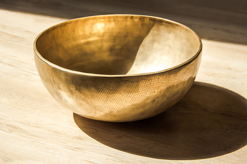 A closeup of a golden round singing bowl in sunlight on a wooden table