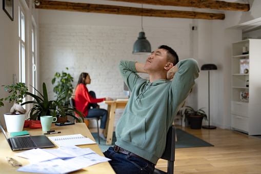 Young Asian man with his eyes closed and hands behind back feeling exhausted while working at home. There is a person in the background.