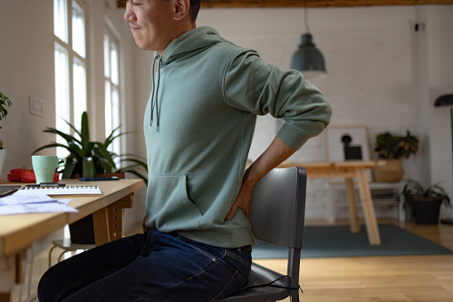 Displeased Chinese man having lower back pain while working at home office.