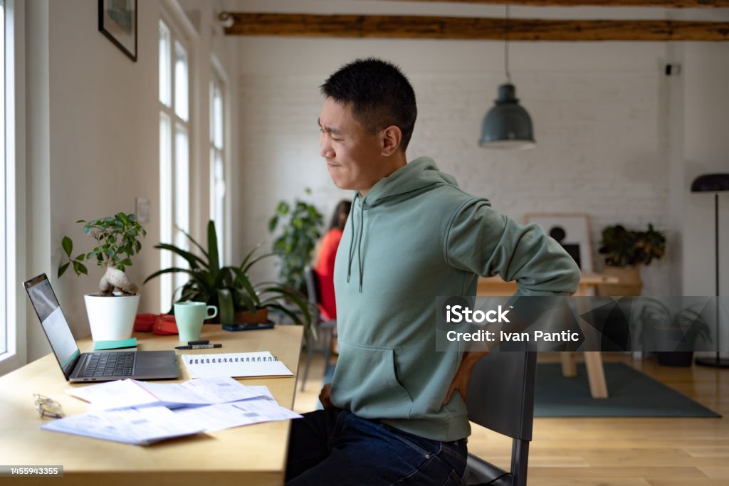 Ugh, my back hurt a lot! Displeased Asian man holding his back in pain while working on a computer at home office. 25-29 Years Stock Photo