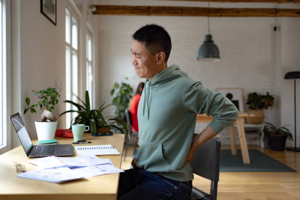 Ugh, my back hurt a lot! Displeased Asian man holding his back in pain while working on a computer at home office. bad posture stock pictures, royalty-free photos & images