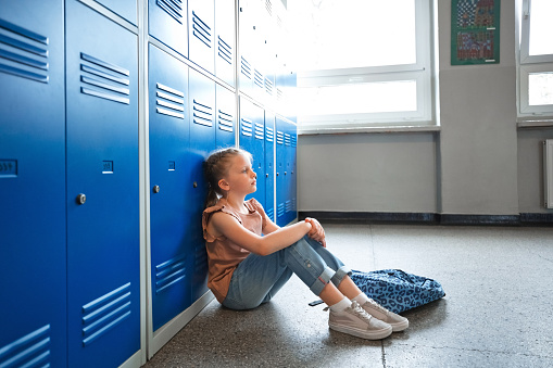 Thoughtful girl sitting on the floor in the school corridor next to lockers, looking away.