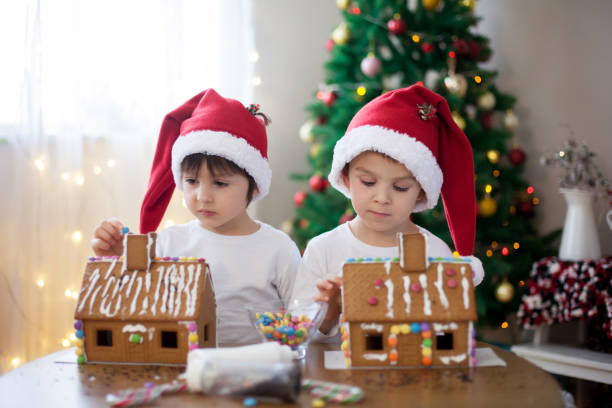 Two sweet boys, brothers, making gingerbread cookies house, decorating at home in front of the Christmas tree, child playing and enjoying, Christmas concept
