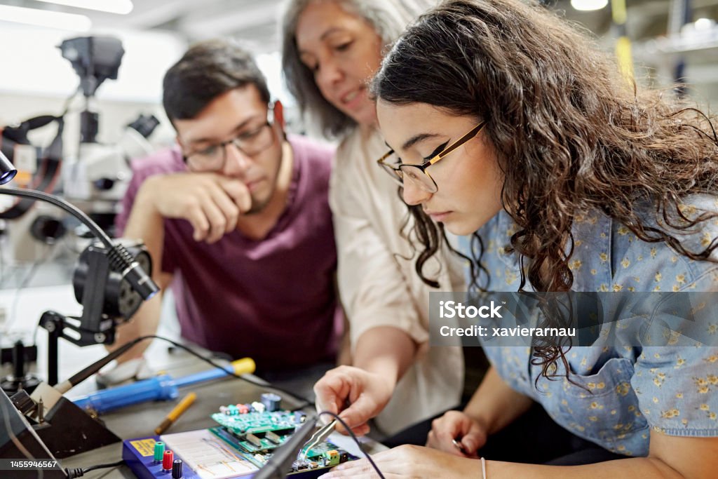 College instructor assisting electrical engineering students Close view of mature female educator standing between early 20s classmates and offering guidance as they work on project. Property release attached. Technology Stock Photo