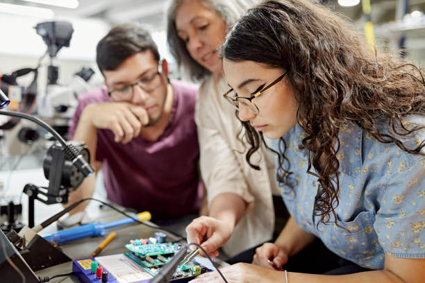 College instructor assisting electrical engineering students Close view of mature female educator standing between early 20s classmates and offering guidance as they work on project. Property release attached. science and technology lab stock pictures, royalty-free photos & images