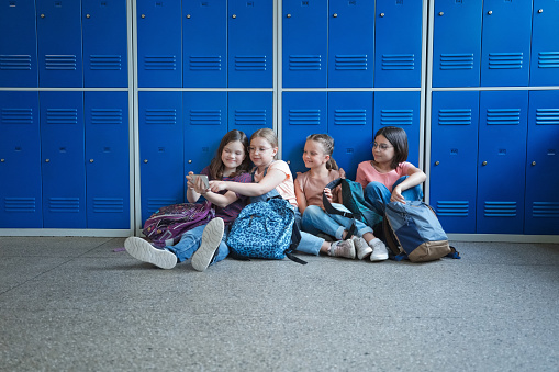 Happy girls sitting on the floor in the school corridor next to lockers and using smart phone.