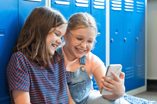 Happy girls sitting on the floor in the elementary school corridor next to lockers and using smart phone.