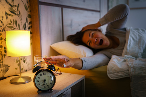 Mature woman yawning in bed early in the morning. And reaching black alarm clock that is on the night table. Selective focus on alarm clock. High resolution 42Mp indoors digital capture taken with SONY A7rII and Zeiss Batis 40mm F2.0 CF lens