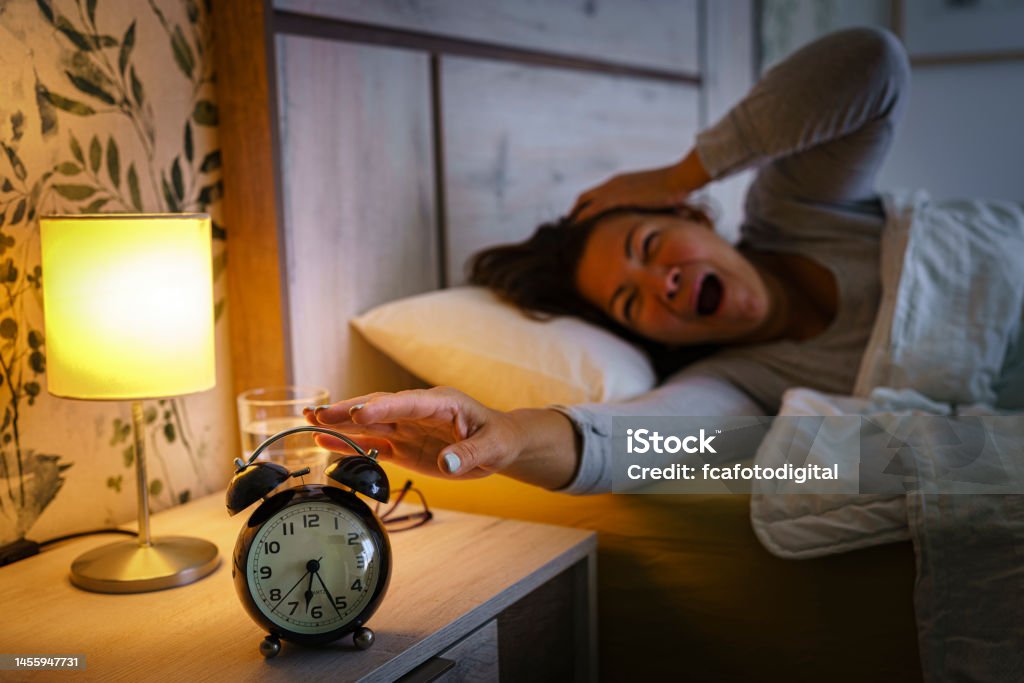 Woman yawning in bed early morning Mature woman yawning in bed early in the morning. And reaching black alarm clock that is on the night table. Selective focus on alarm clock. High resolution 42Mp indoors digital capture taken with SONY A7rII and Zeiss Batis 40mm F2.0 CF lens 50-54 Years Stock Photo