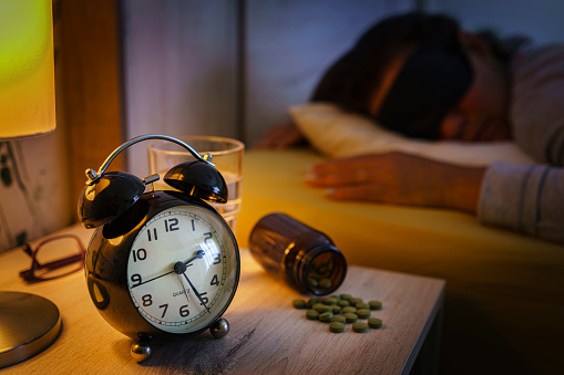 Sleep disorder: close up of a night table with alarm clock and pill bottle. A sleeping woman wearing sleeping mask is out of focus at background. High resolution 42Mp indoors digital capture taken with SONY A7rII and Zeiss Batis 40mm F2.0 CF lens