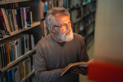 Senior Caucasian male with gray hair and a beard reading a book. Standing between the bookshelves in a library wearing eyeglases.
