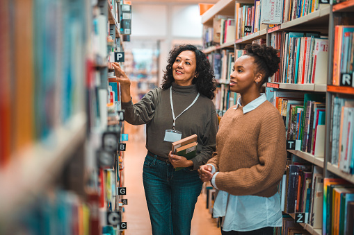 Mid adult Hispanic librarian escorting a student in a library. Searching for a book on shelve.