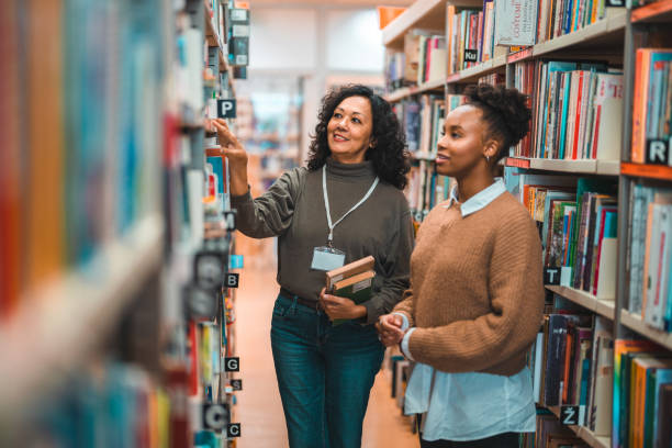 средний взрослый латиноамериканский библиотекарь сопровождает студента в библиотеке - back to school young women cheerful happiness стоковые фото и изображения