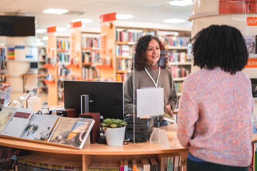 Mid adult Hispanic female librarian serving a costumer in a library. Helping with the decision on what book to borrow.