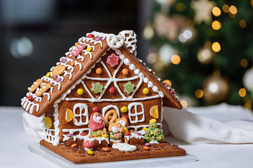 Christmas house made from gingerbread. Playfully decorated with colourful edible figures. Christmas tree in background with lights. Festive New year atmosphere and mood.