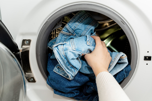 Person putting jeans into the drum of a washing machine, front view. Washing dirty jeans in the washer
