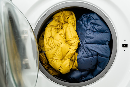 Blue and yellow winter puffer jacket in the drum of washing machine in laundry room. Washing dirty down jacket in the washer