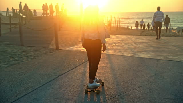 Young woman with brown hair skating on her skateboard on a boardwalk during sunset,rear view,sunbeam