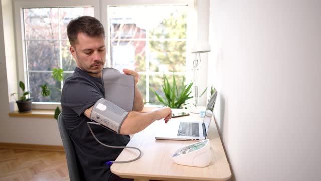 Caucasian man, adjusting blood pressure gauge, while working on laptop from home office