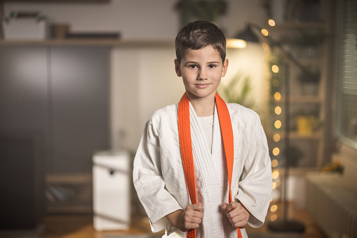 Portrait of a cute young boy in white kimono with orange belt around his neck standing in living room at home.