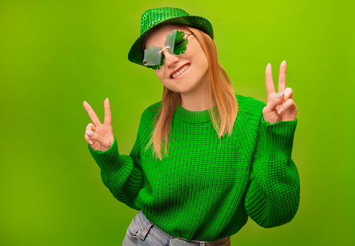 St Patrick Day celebration concept: happy smiling young woman in green sweater, clover shaped glasses with hat showing peace gesture v sign isolated on colorful green background.\n\nCelebrating national irish holiday.
