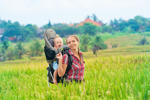 Nature walk in green rice terrace field. Happy mother hold little traveller in carrying backpack. Baby ride on woman back. Travel adventure, hiking with child carrier, family summer vacation on Bali.
