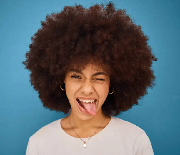 Face, portrait and woman with tongue out being silly and goofy on a blue studio background. Amusing, comic and crazy african american female making a funny, humour face on a backdrop
