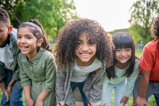 A small group of diverse children stand side-by-side outdoors in a park as they pose for a portrait.  They are each dressed casually and are smiling as they lean into the camera with their hands on their knees.