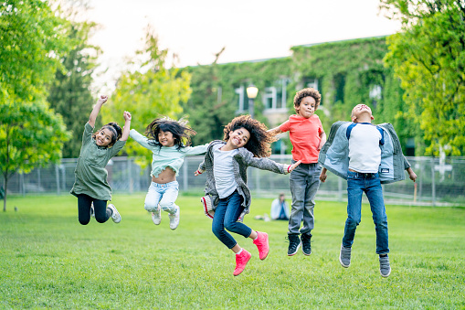 A small group of school aged children run through the grass while spending time at a local park.  They are each dressed warmly in layers and laughing as they play joyfully.