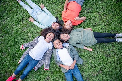 A small group of school aged children lay in a circle in the grass with their heads together as they pose for a portrait.  They are each dressed casually and are smiling as they enjoy the fresh air.