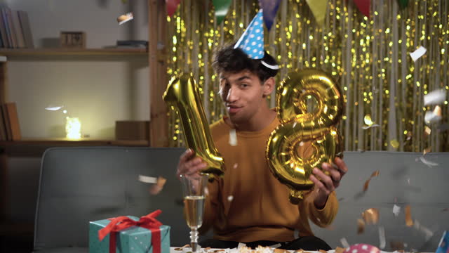 A birthday guy on his 18th birthday with gold number balloons. Excited eighteen arabic man celebrating and having fun at home. student on happy birthday.