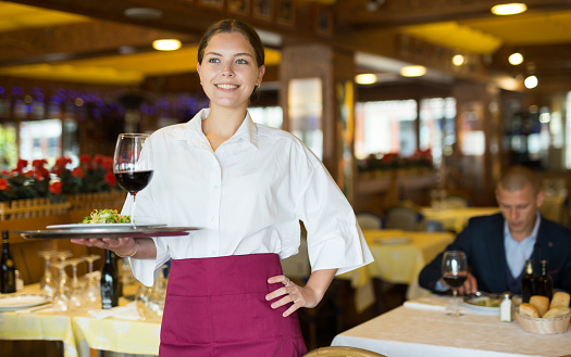 Smiling woman waiter with serving tray carrying order for visitors in restaurant