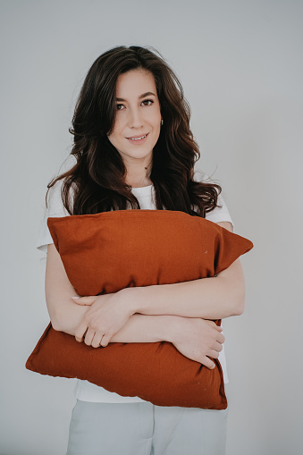 Vertical shoot of brunette young adult caucasian woman standing at home, toothy smiling, hugging pillow, looking at camera. Attractive hispanic female at weekend relaxing. Healthy, successful people.