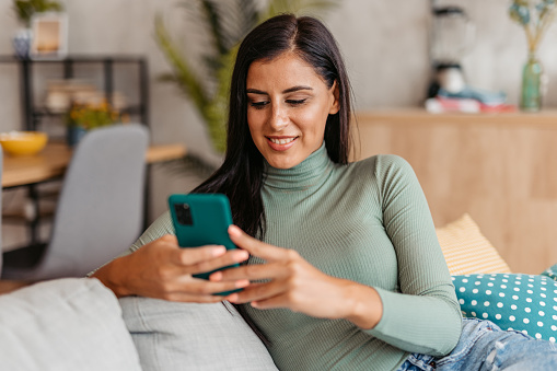 Smiling young beautiful woman relaxing at home, texting on smartphone.