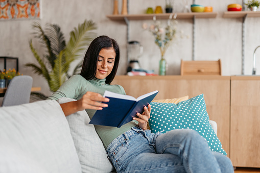 Beautiful young woman sitting on the sofa at home and reading a book.