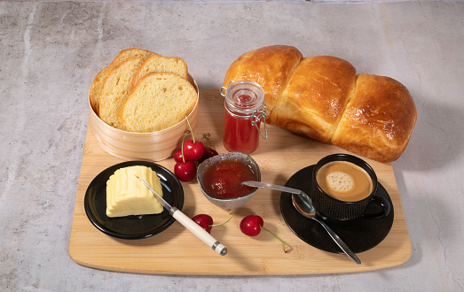 French breakfast with brioche and cherry jam, butter and coffee with foam served on a cutout wooden tray. A few fresh cherries are placed on the tray