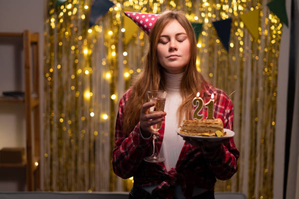 caucasian girl makes a wish closing her eyes holding a birthday cake with candles number 21 in her hands - 21e verjaardag stockfoto's en -beelden