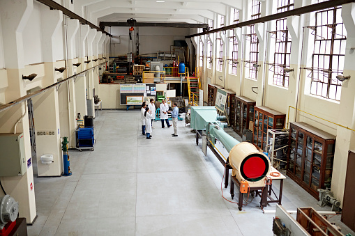 High angle view of young men and women in lab coats standing in large cement work area and listening to male educator. Property release attached.