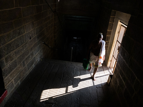 Trichy, Tamil Nadu, India - February 2020: An elderly Indian temple priest walking in a dark passage of the Rock Fort temple lit only by sunlight from a door.