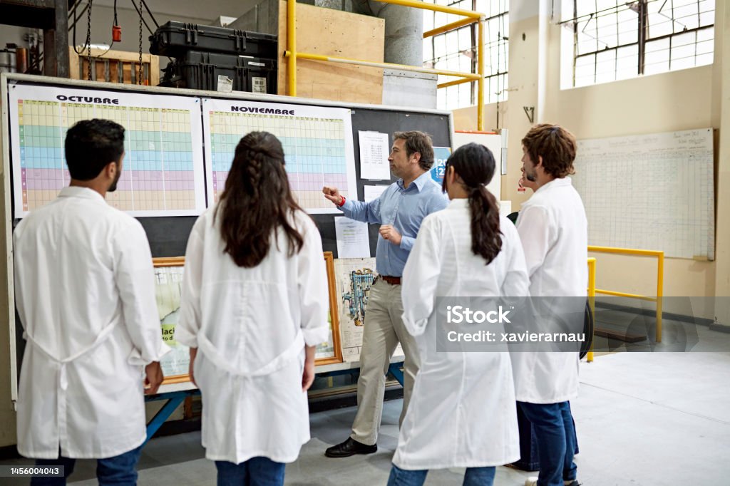 College instructor discussing curriculum plans with students Young men and women in lab coats standing and listening to mature male educator describe assignments for the term. Property release attached. Education Training Class Stock Photo