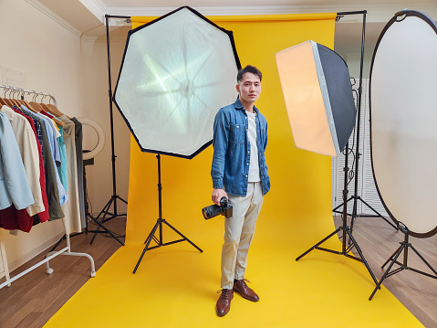 asian young male photographer holding professional camera for shooting standing in front of yellow background at studio