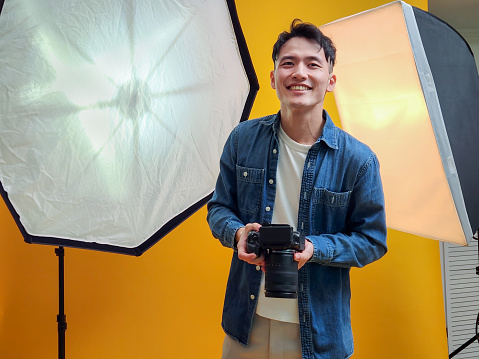 portrait asian young male photographer holding professional camera for shooting standing in front of yellow background at studio