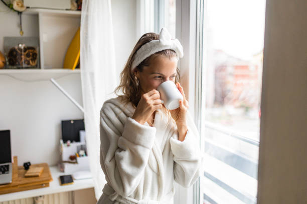 Young woman at home drinking coffee looking through window stock photo