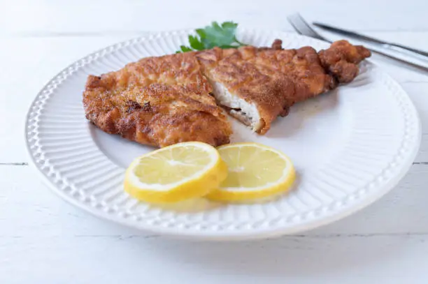 Delicious homemade pan fried chicken schnitzel. Served on white plate with garnish such as sliced lemon and parley isolated on white wooden table background. Closeup and front view with cross section view.