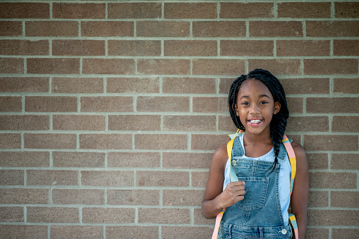 A female Elementary student of African decent, poses with her backpack for a portrait outside.  She is dressed casually and is smiling as she enjoys the fresh air.