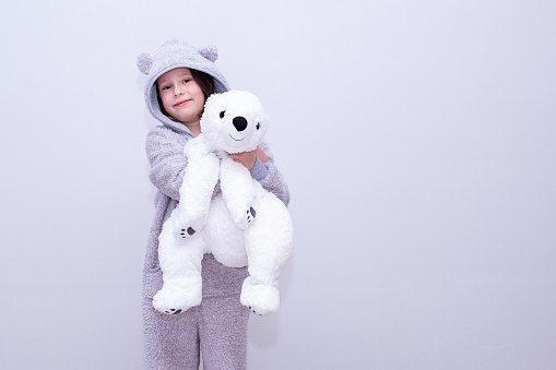 Portrait of a Caucasian girl 7 years old in warm home clothes stands against the wall and holds a soft toy, a bear. Smiling and looking at the camera.
