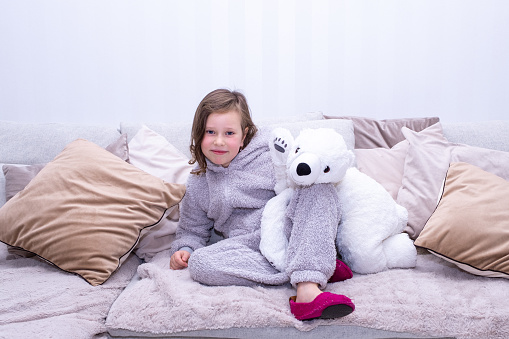 Portrait of a Caucasian girl 7 years old in warm home clothes sits on the sofa, smiles happily and hugs a soft toy, a polar bear.