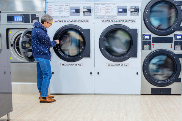 A close-up caption from a diagonal view of an opened, metallic lustered clothes washer’s door in a public launderette. The background is out of focus, daylight, no humans. The focus is on the washing machines opened door. Horizontal taken picture with a full frame camera and a prime lens on daylight.