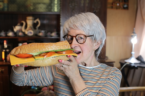 A senior woman in her sixties is eating an extra large sandwich. She has short gray hair and wears eyeglasses.