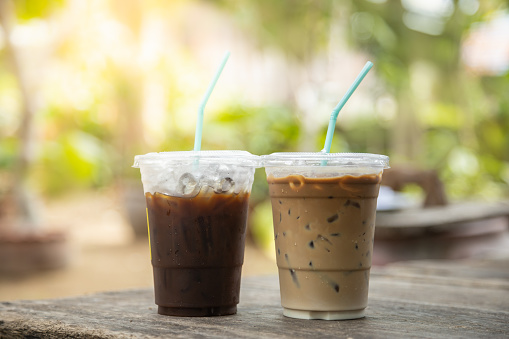 Closeup of takeaway plastic cup of iced black coffee Americano and coffee latte on wooden table with green nature background.